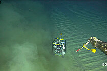 Aufnahme vom Meeresgrund, ein kleines Gestell mit Sensoren steht auf dem Grund, der Arm eines Unterwasserroboters ragt ins Bild, von links kommt eine Sedimentwolke ins Bild