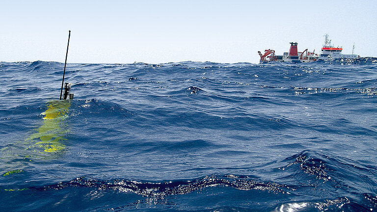 Eine gelbe Messboje im Wasser, am Horizont ein Forschungsschiff